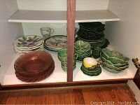 Cabinet shelf with pink glass plates, and green majolica cabbage leaf ceramic dishes including bowls and plates, plus some cups.