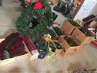 Photo showing two artificial Christmas wreaths with red berries, wreath stands, multiple boxes and baskets filled with multi-colored Christmas string lights, red mesh tree skirt, and other decorative holiday items spread on floor.