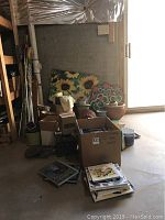 Wide view showing cluster of assorted ceramic and wicker planters, outdoor garden lamps including one with sunflower design, boxes and stacks of nature books on the floor.