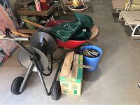 Overall wide view of wheelbarrow, garden hose caddy, weed eater box, blue bucket with gloves and small tools