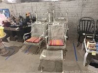 Photo showing six white metal outdoor chairs with four having striped cushions. Chairs have decorative backs and armrests. The set is arranged on concrete floor.