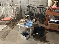 White metal outdoor furniture set including a bench and a table partially visible, two black metal outdoor chairs with curved armrests and vertical slat backs, and various other items including boxes and a wooden workbench.