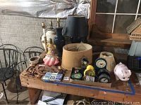Overview of the lot showing table clocks, piggy bank, lamps with shades, porcelain doll, and other small decorative items arranged on a wooden table.