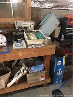 Singer sewing machine on wooden workbench with teal sewing scissors, thread bobbin case, sewing accessories, vacuum bags, and fabric/boxes under bench.
