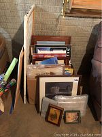 Photo showing a variety of framed prints and posters stacked vertically, including some larger white canvases and wooden framed art