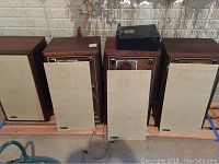 Four Advent Loudspeaker cabinets with two speaker stands on wooden boards in storage area. Three grills removed from front panels.