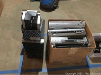 Box containing multiple silver DVD and VHS recorders and players next to Sony subwoofer and box of satellite speakers.