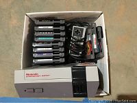 Top-down photo showing the NES console, one rectangular NES controller, and 12 NES game cartridges in a box.