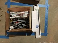 View of Wii console with game cases, cables, and sensor bar in a box on the floor.