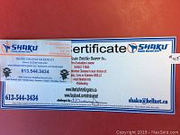 Photo of the printed Shaku Family Martial Arts gift certificate and a business card from the instructor, Sensei Helena Greenley. The certificate outlines the included services: a pre-evaluation lesson, a Shaku T-shirt, and one month of unlimited classes in various martial arts programs. The certificate has branding in blue and orange.