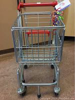 Front view of the mini metal shopping trolley for children, showing metal wire basket, red handle and four caster wheels.