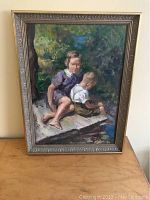 Full view of framed oil painting on panel showing two children sitting near water with dense foliage background.