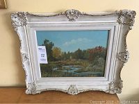 Frontal image of the framed landscape painting showing the entire artwork and ornate white frame with decorative corner elements and wear.