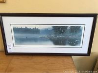 Framed print showing the full artwork including frame and matting, positioned on a wooden surface against a wall.