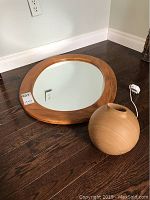 Oval mirror with wooden frame resting on floor, reflecting wall and outlet.