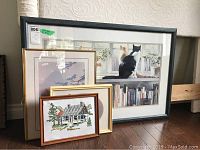 Six framed pieces of art and needlework stacked against a wall, largest shows a cat on a windowsill, smaller framed cross-stitch in front.