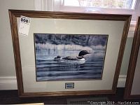 Framed print featuring a loon on a lake with a wooden frame and matting, showing the overall artwork and setting.