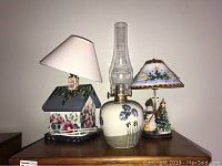 Photo of two electric lamps and one ceramic antique oil lamp with floral designs and a glass chimney on a wooden surface against a plain wall.