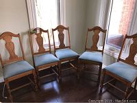 Five wooden high back dining chairs with carved backs and light blue upholstered seats, arranged in a semicircle near a window on wooden floor.