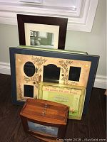 Photo showing several photo frames stacked against a wall near a window, including a dark brown frame and a large collage frame with nature motifs, along with photo albums and a small wooden box.
