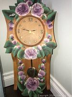 Wooden wall clock with painted purple flowers and green leaves, round clock face with black numerals and gold hands, pendulum showing below.