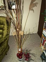 Full view of tall brown pail filled with dried grasses, curly twigs, floral pods, artificial red apples at base and loose dried floral on the floor near bookshelf and green chair.