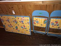 Photo of children's folding table and two folding chairs leaning against wood-paneled wall. Shows blue metal frame, yellow plaid surface with clown pattern. Visible paint finish wear and stains.