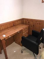Pine wood desk with turned legs, three drawers, and some surface wear, next to a black rolling office chair with visible damage.