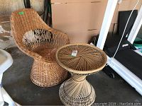 Wicker chair and matching wicker table shown from frontal angle on concrete floor with surrounding items in background.