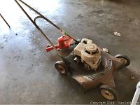 Wide view of the full lawn mower showing rusty metal deck and orange electric hedge trimmer on top of it.