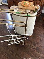 White vinyl laundry hamper, brass blanket rack, white metal and plastic hanging rack shown together with some fabric items on the hamper