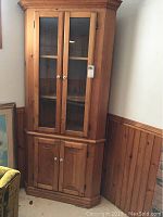 Front view of the wooden corner cabinet showing top glass doors and bottom wooden doors with white round knobs, cabinet situated in a corner.