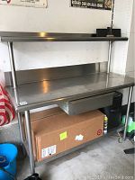 Stainless steel prep table with top shelf, drawer, and lower shelf with visible wear and scratches.