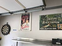 Wide shot showing the dart board mounted on a wall next to two vintage movie posters: Casablanca and Creature from the Black Lagoon.