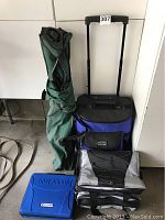 Green canvas folding beach chair, blue soft-sided rolling cooler bag with telescoping handle, blue plastic first aid kit, and blue/black cooler bag with grip handles together.