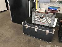 Black metal trunk closed, with milk crate of books on top, showing size and condition of trunk