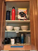 Full cabinet shelves showing all items including waffle maker box, water flasks, mugs, bowls, and tumbler