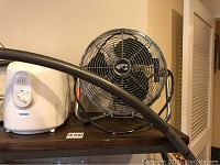 Side view of the white Vicks humidifier and chrome Hampton Bay metal fan side by side on a wooden shelf.