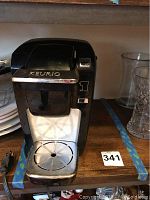Front view of black Keurig coffee maker on wooden shelf, shows power cord and drip tray with circular pattern.