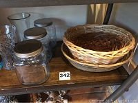 Shelf showing three round woven baskets and three clear glass storage canisters with metal lids