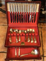 Opened wood flatware chest displaying mixed pattern silver plated knives, forks, spoons, and serving pieces arranged in lined compartments.