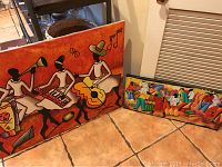 Both canvas paintings placed on a tiled floor near a metal stand and vent, showing size contrast and detail.