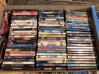 Wide shot of a couch with a large stack of DVDs arranged vertically showing titles. The DVDs are mostly children and family movies.