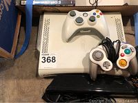 White Xbox 360 console with two wireless controllers and a Nintendo GameCube controller on top and a sensor bar behind