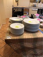 Multiple stacks of white Mikasa dishware pieces including dinner plates, bread plates, soup bowls, and others arranged on a table with a plaid tablecloth.