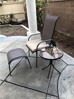 Outdoor patio set with chair, footstool, and glass top table arranged on concrete patio with brick wall background