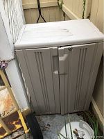 Beige heavy plastic storage cabinet with two doors, closed, showing dirt on surface, next to yellow metal hand truck with black wheels.