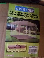 Packaging box photo showing image of assembled gazebo, brand Arctic Sky, size 10x10 feet, with mosquito netting and beige color. Mentions carry bag with wheels.