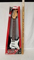 Full view of toy guitar in packaging with measuring stick to the side showing length about 24 inches.