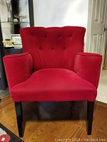 Front view of a red velvet tufted armchair with padded armrests and black wooden legs on hardwood floor.
